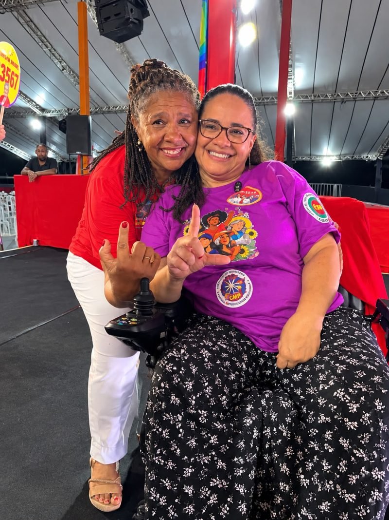 Ana Campos, cadeirante, e Marta Rodrigues aparecem sorrindo e abraçadas em um evento. Ana veste camiseta roxa e faz um gesto com o dedo indicador levantado, enquanto Marta, de camisa vermelha e calça branca, faz um gesto de resistência.